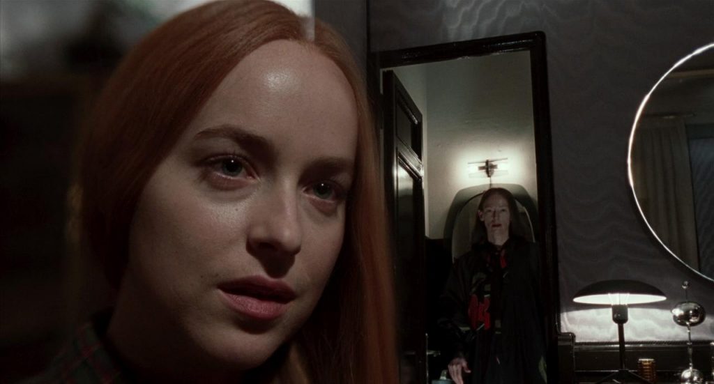 A shot from the movie Suspiria. Dakota Johnson is close up on the left side, while a mirror behind her shows Tilda Swinton ont he right side of the photo